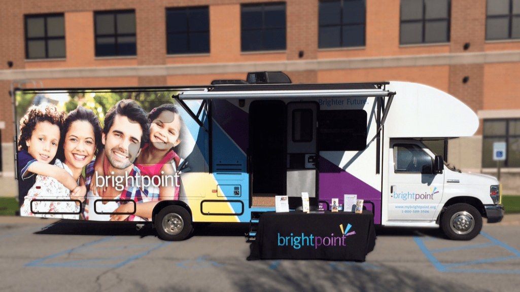 Recreational Vehicle with a Brightpoint branded wrap and photo of a smiling family on the exterior. An outreach table with a branded tablecloth sits in front of the RV